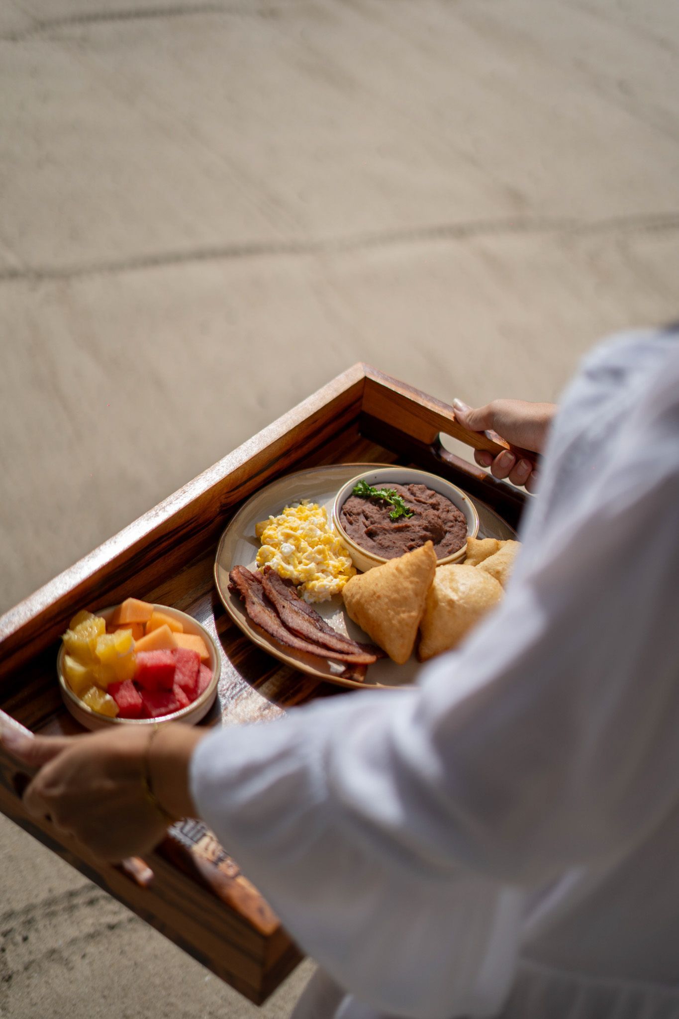 A person carrying a wooden tray with a plated breakfast and fruit on a stone floor