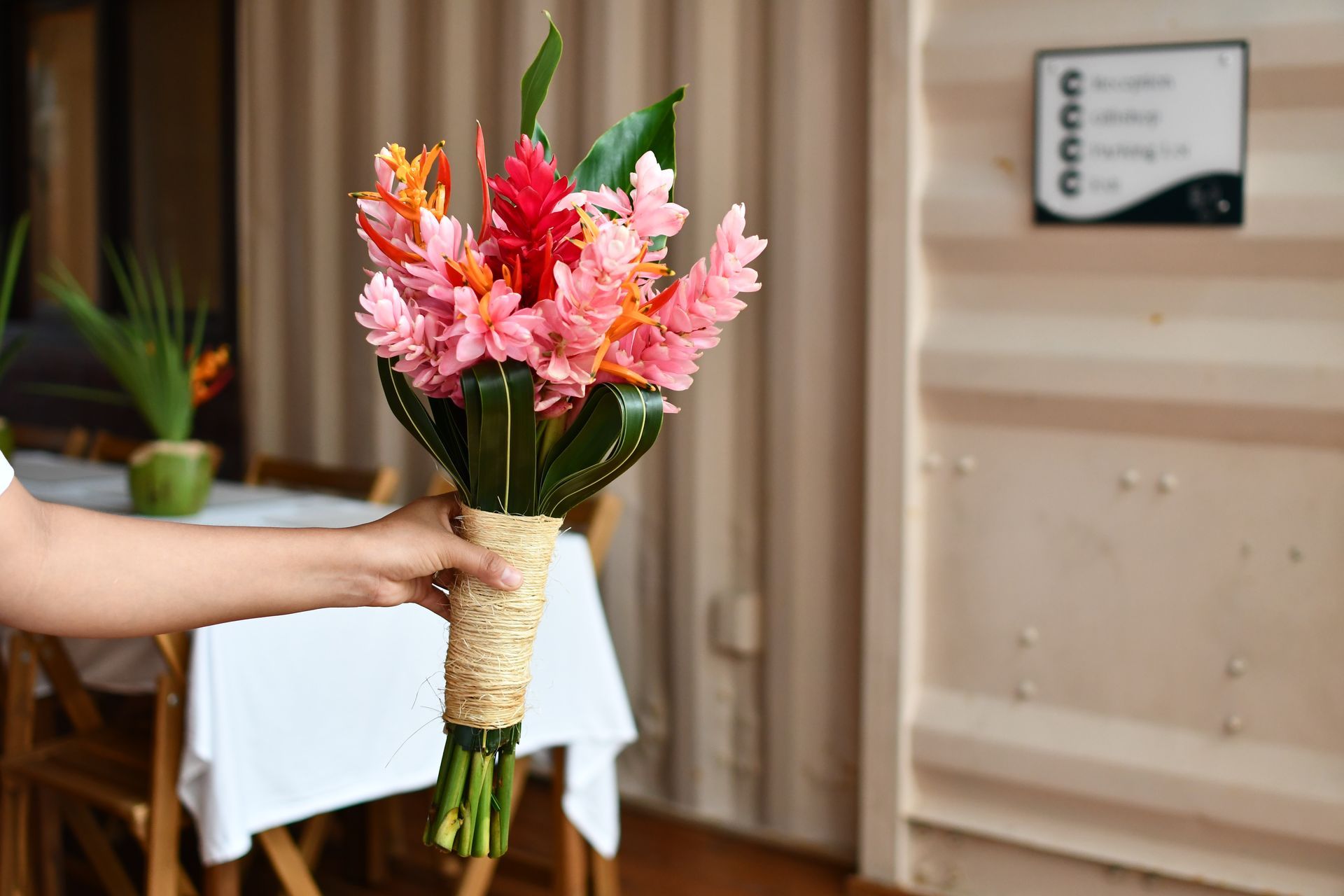 Hand holding a bouquet of pink flowers in a wrapped stem, indoors beside a table and door