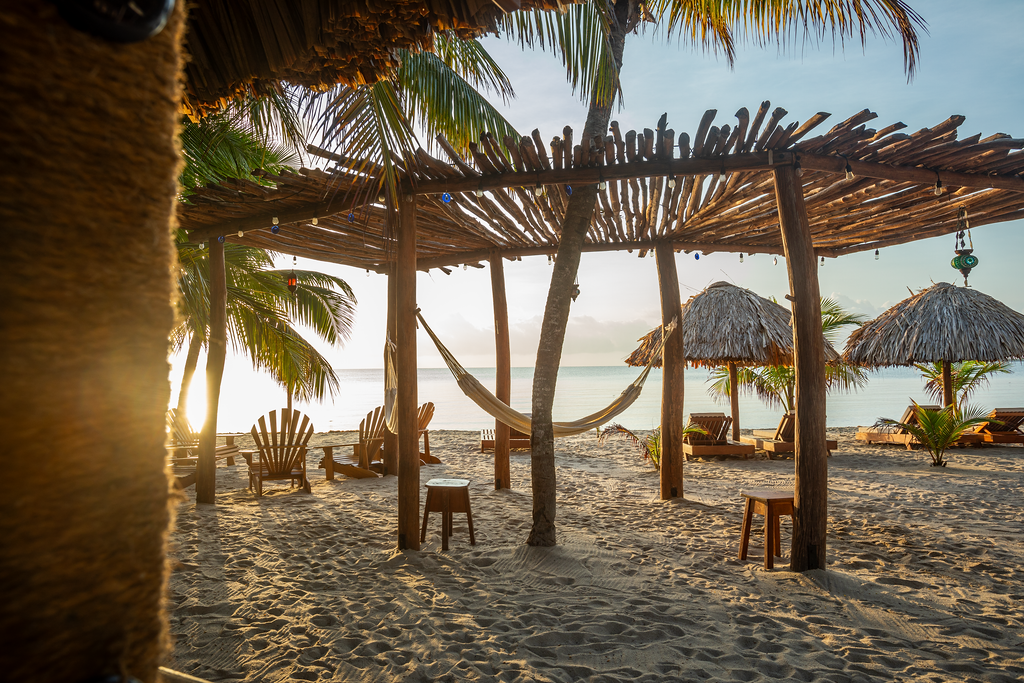 Sunset beach scene with thatched huts, wooden pergola, palm trees, and chairs on the sand