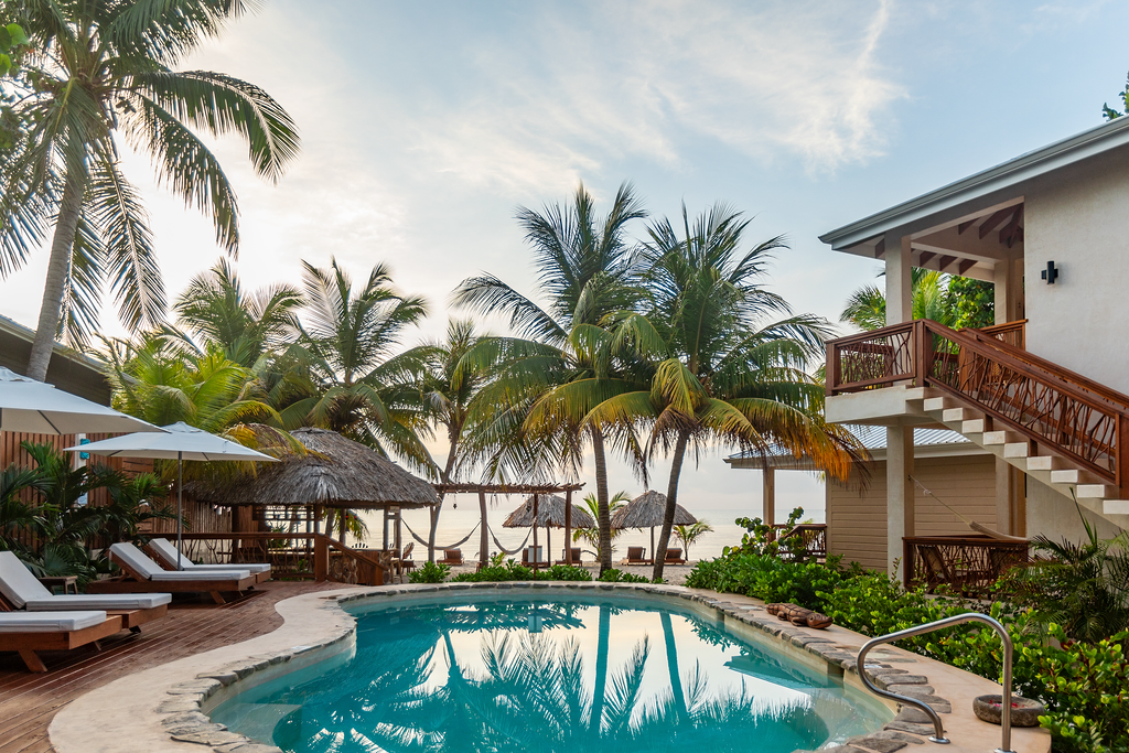 Tropical resort pool with palm trees, lounge chairs, and a villa overlooking the water at sunset