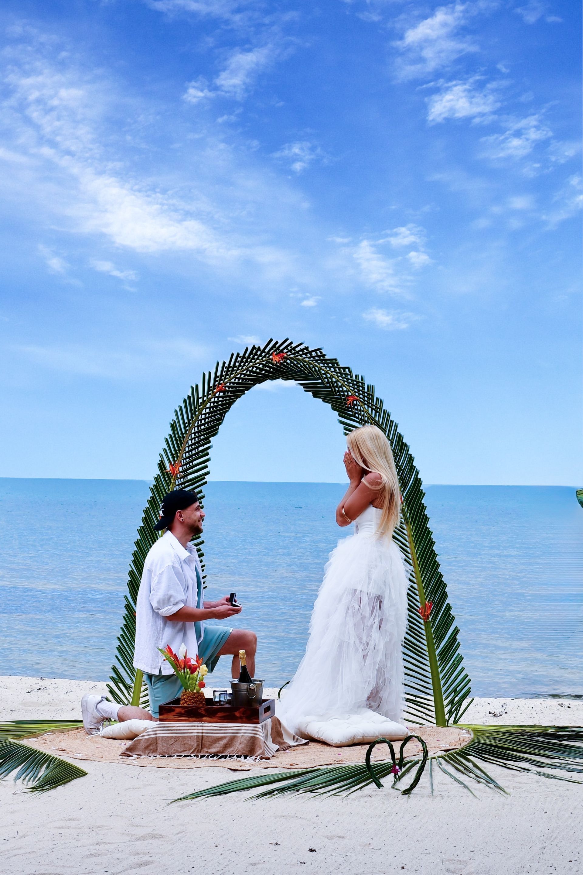Beach wedding ceremony under leafy arch by the sea, with bride in white dress and groom kneeling nearby