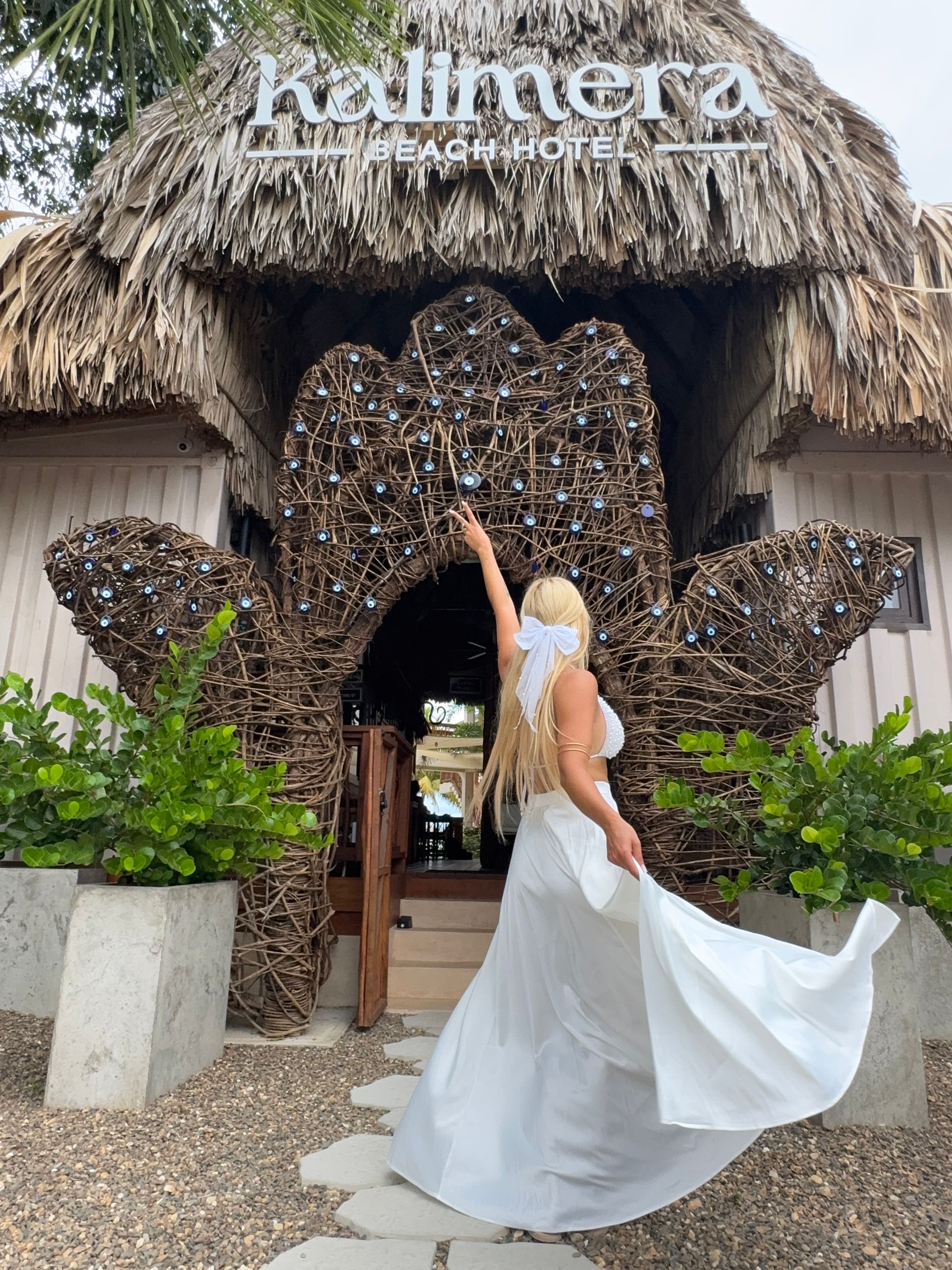 Woman in white dress poses before a thatched resort entrance with a sign overhead