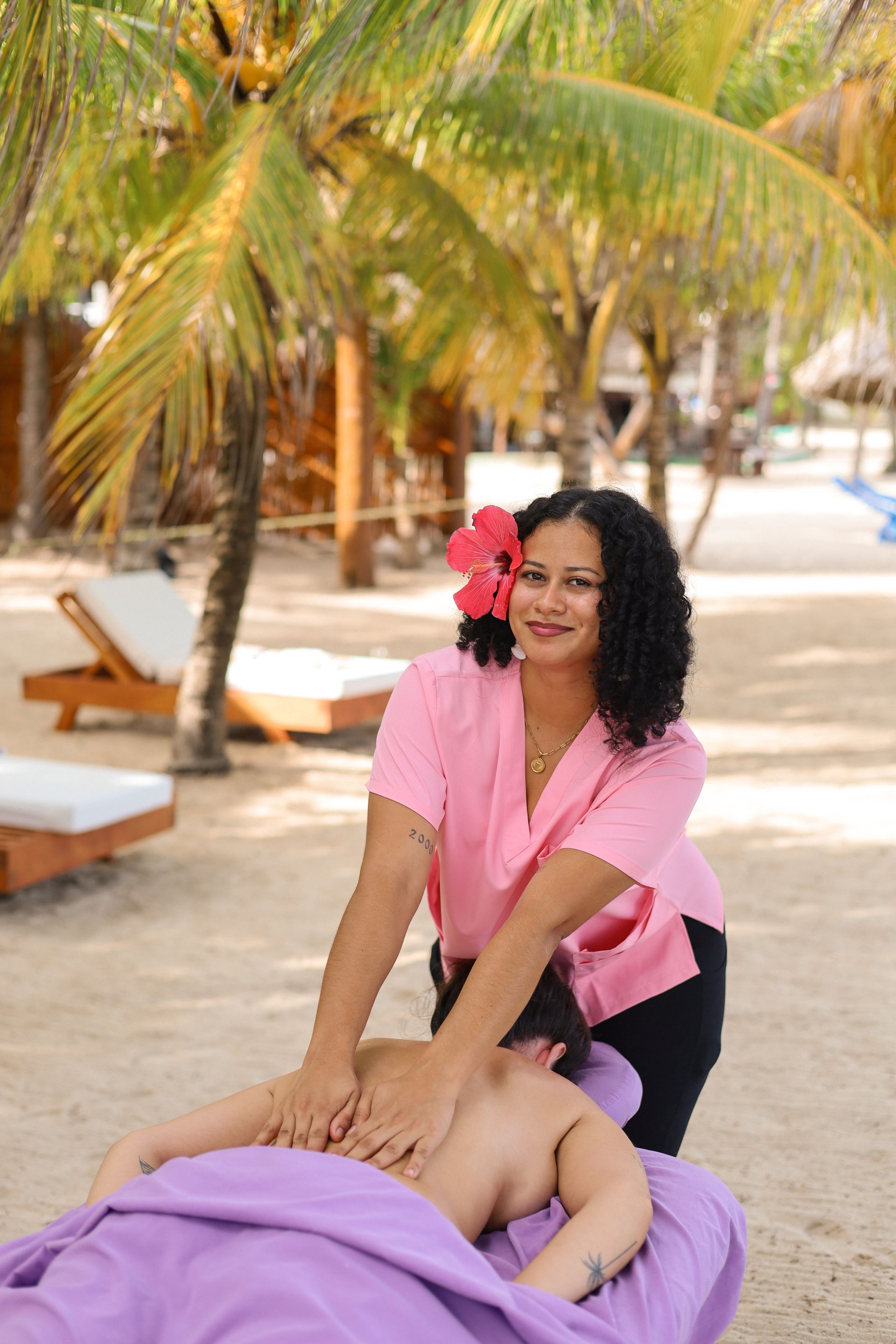 Massage therapist giving a back massage outdoors under palm trees, smiling in a pink shirt.
