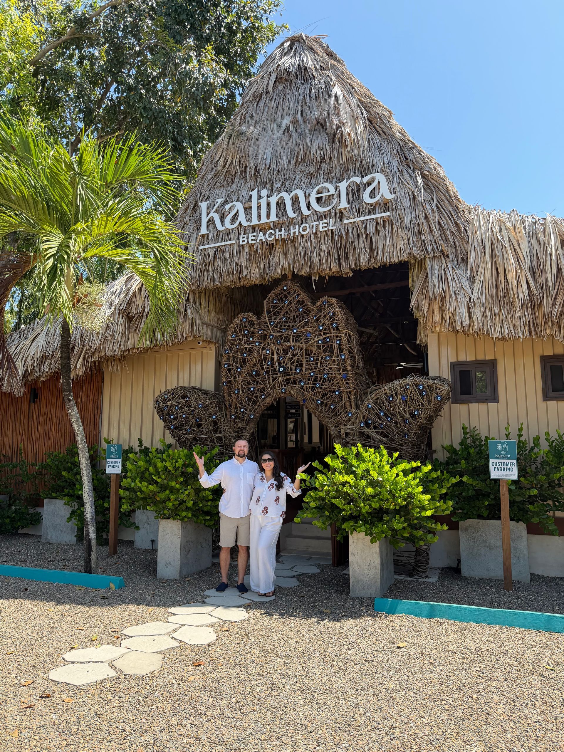 Two people posing under a thatched Kalimera sign at a tropical resort entrance