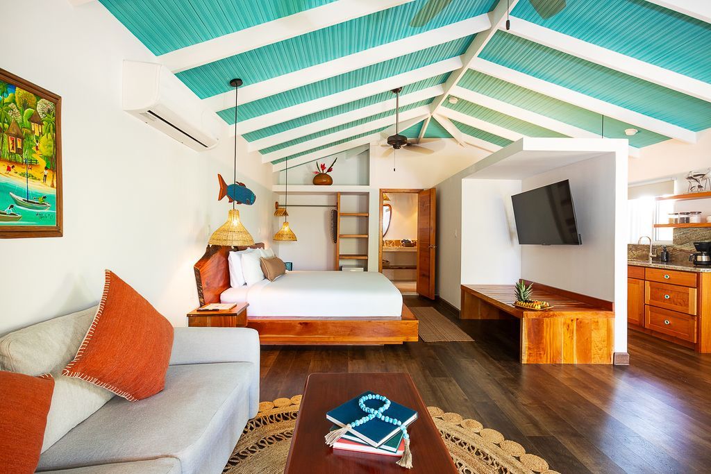 Bright tropical bedroom with teal ceiling beams, bed, sofa, TV, and wooden furniture
