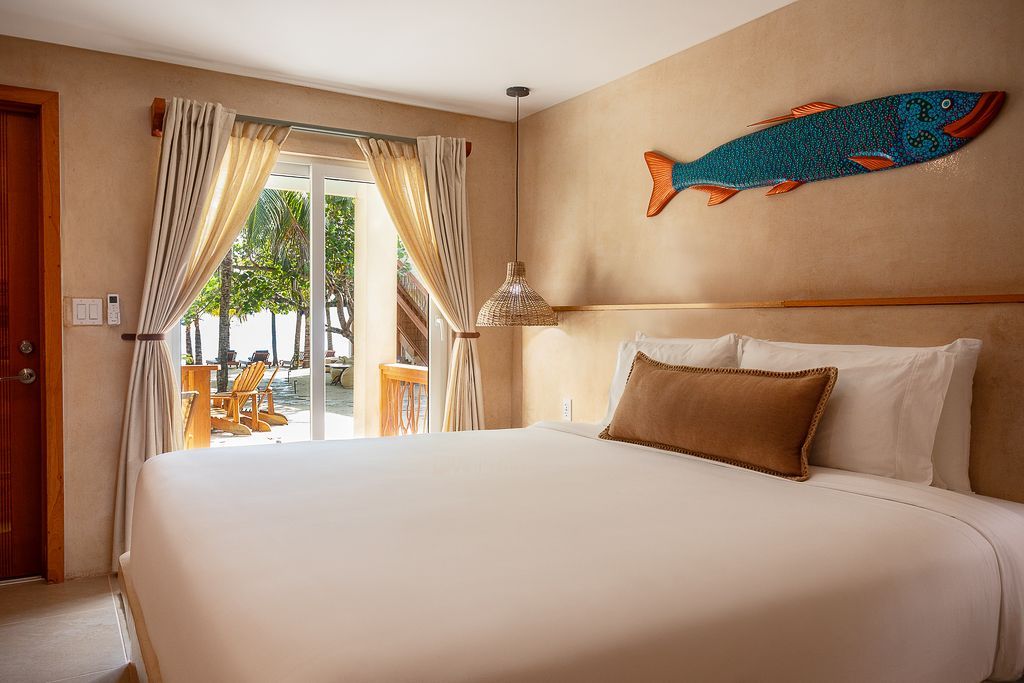 Bright hotel bedroom with large bed, fish wall art, and open doors to a sunny balcony.