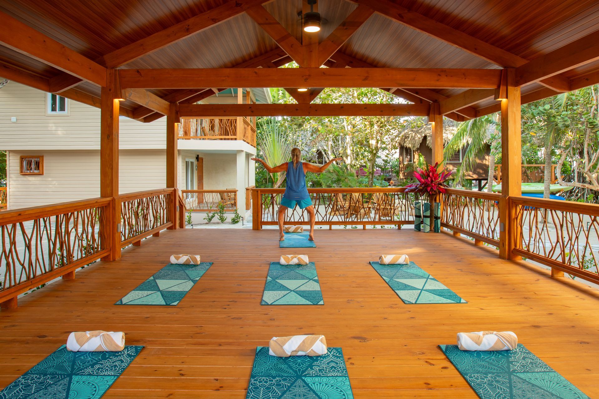 Wooden yoga pavilion with teal mats and towels, a person standing at the front, surrounded by greenery.