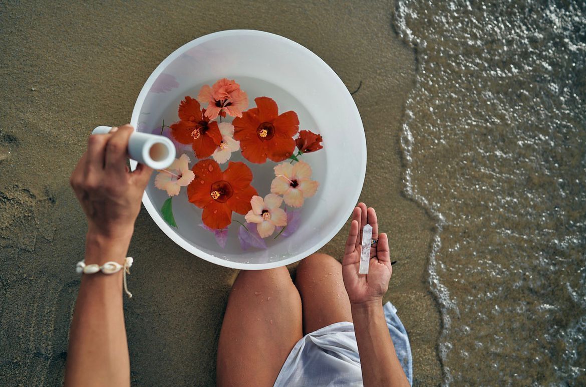 Hands pouring water over colorful flowers in a white bowl on a sandy beach