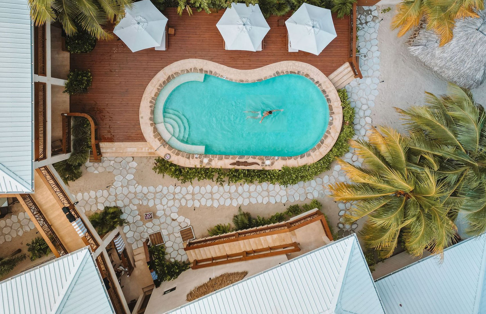 Aerial view of a turquoise pool with white lounge chairs, palm trees, and wooden decking in a resort courtyard