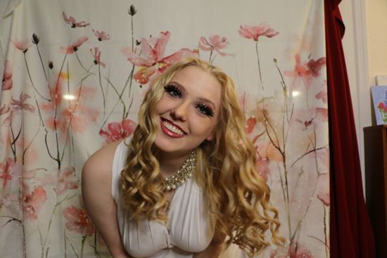 A woman in a white dress is smiling in front of a floral curtain.