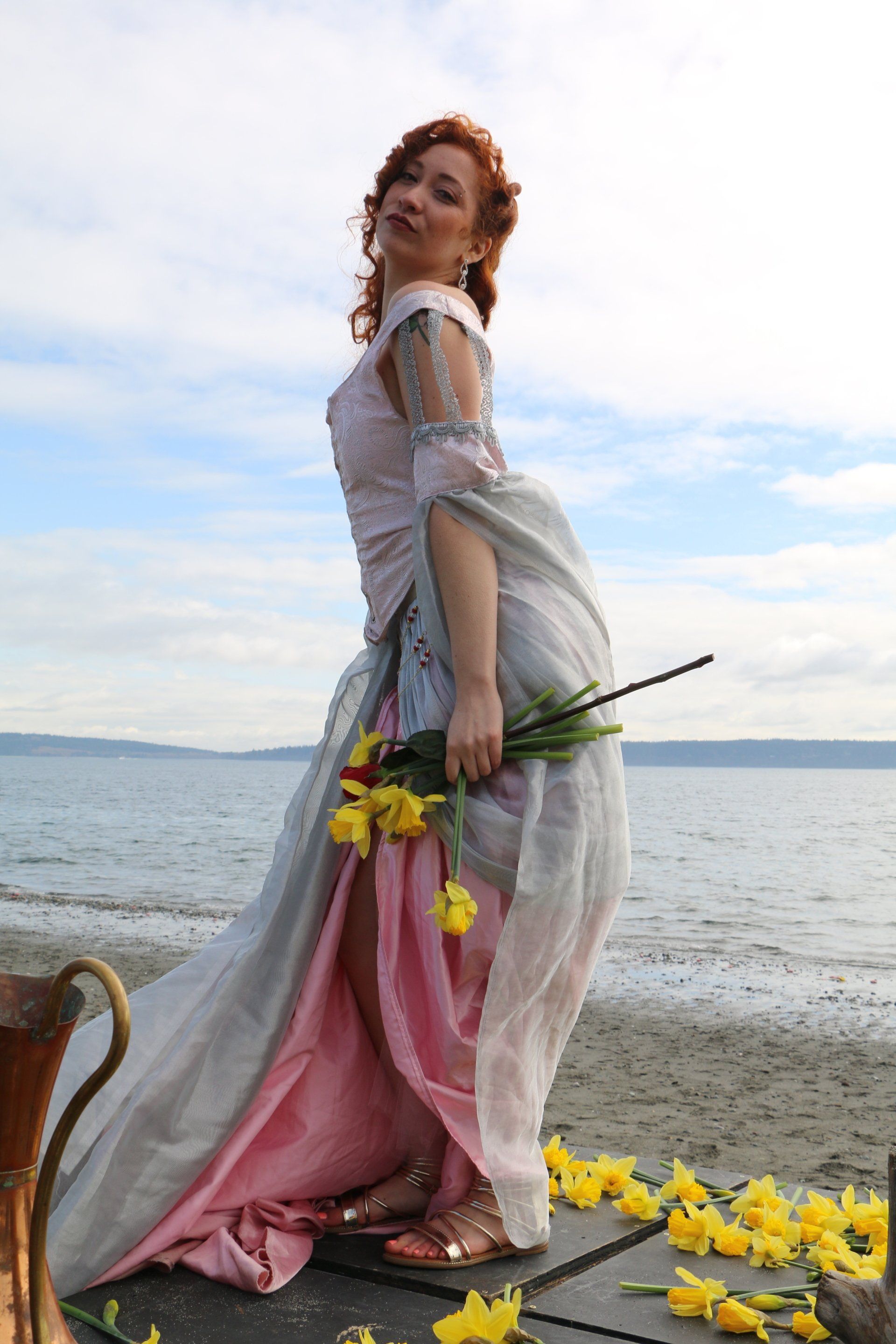 A woman in a long dress is standing on a beach holding flowers.