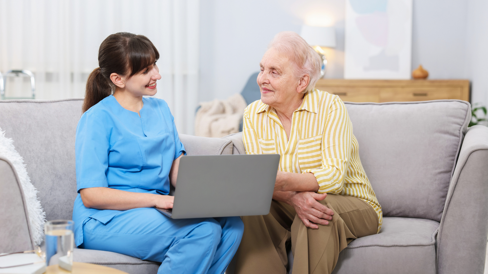 Caregiver showing laptop to senior on a couch, indoors. They both are smiling.