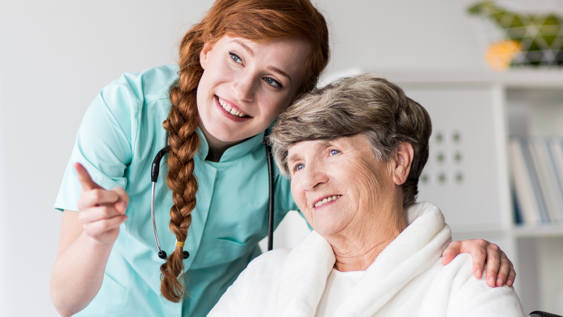 Nurse pointing, arm around elderly woman in bathrobe; indoors, cheerful expressions.
