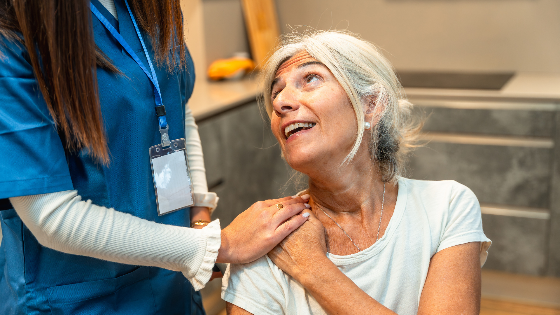 Nurse comforts an older woman, hand on her shoulder, both looking up with smiling expressions in a kitchen setting.
