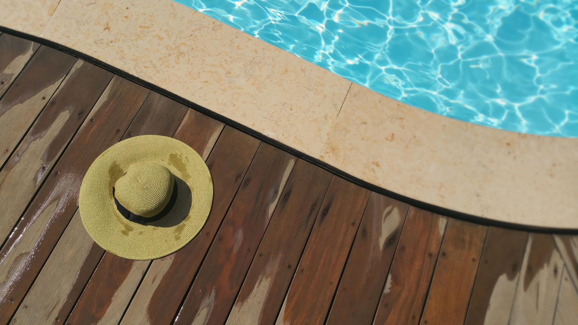 A straw sun hat rests on a wooden deck next to the curved edge of a bright blue swimming pool.