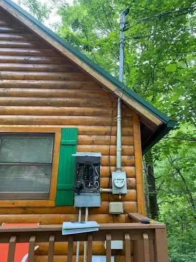 A wooden cabin with electrical equipment attached to the exterior. Green shutters and trim, set in a wooded area.