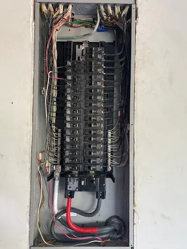 Electrical panel with numerous wires and circuit breakers, installed in a wall cavity.