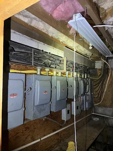 Row of gray electrical meters mounted on a wooden wall, with wiring and overhead lighting.