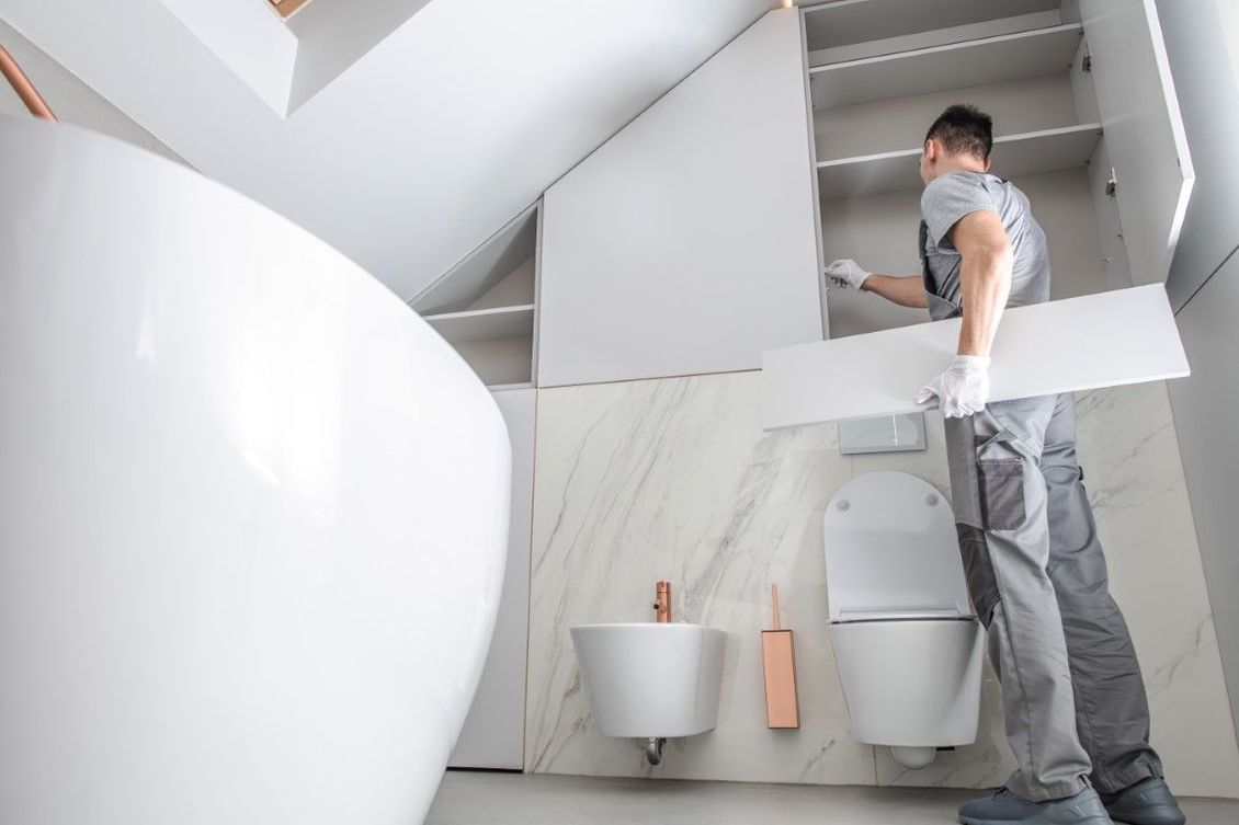 Worker installs an upper bathroom cabinet above a wall mounted toilet and sink in a modern bathroom.