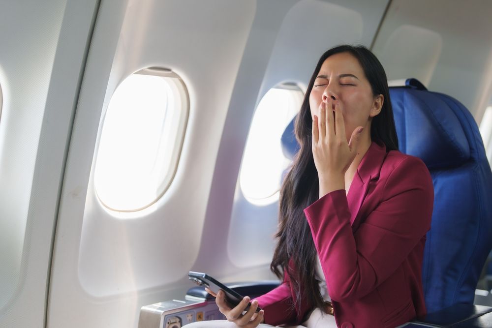 Passenger yawning on an airplane seat, holding a phone and covering her mouth.
