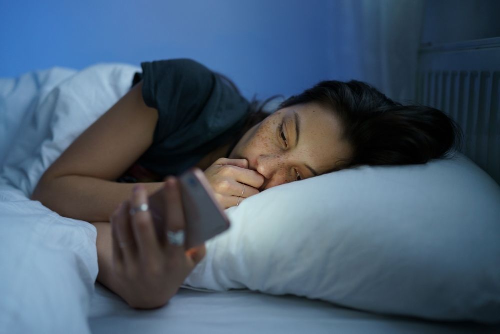Person sleeping in bed while holding a smartphone, with a pillow and white bedding.