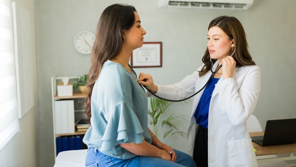 A healthcare professional in a white coat uses a stethoscope to examine a patient in an office setting.