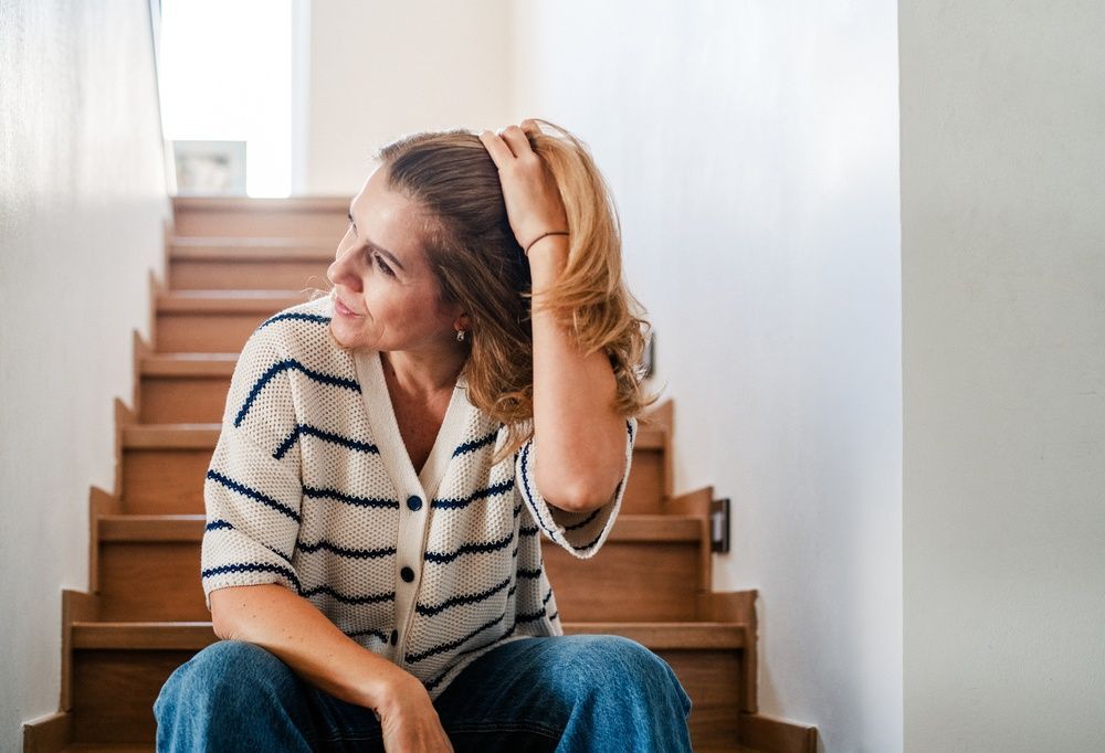 Woman sitting on wooden stairs, touching her hair and looking to the side in a bright room.