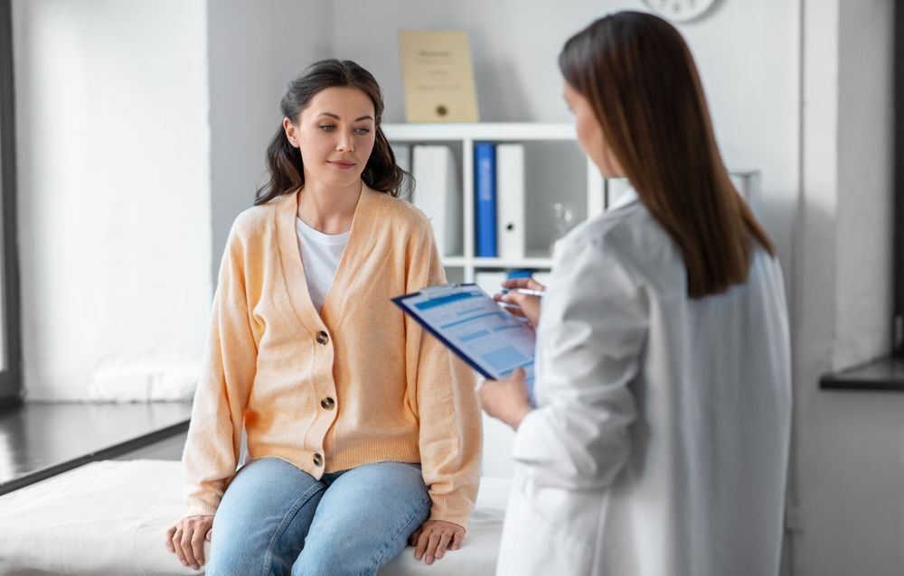 Doctor consulting with a seated patient in a bright clinic office, holding a tablet.