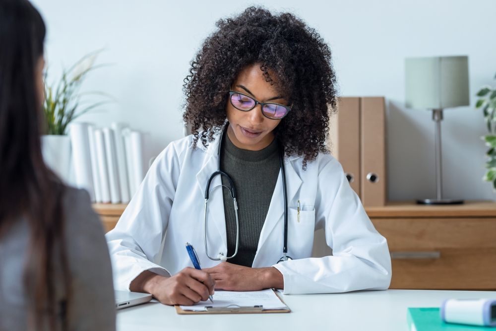 Doctor writing on a clipboard at a desk while consulting with a patient in an office.