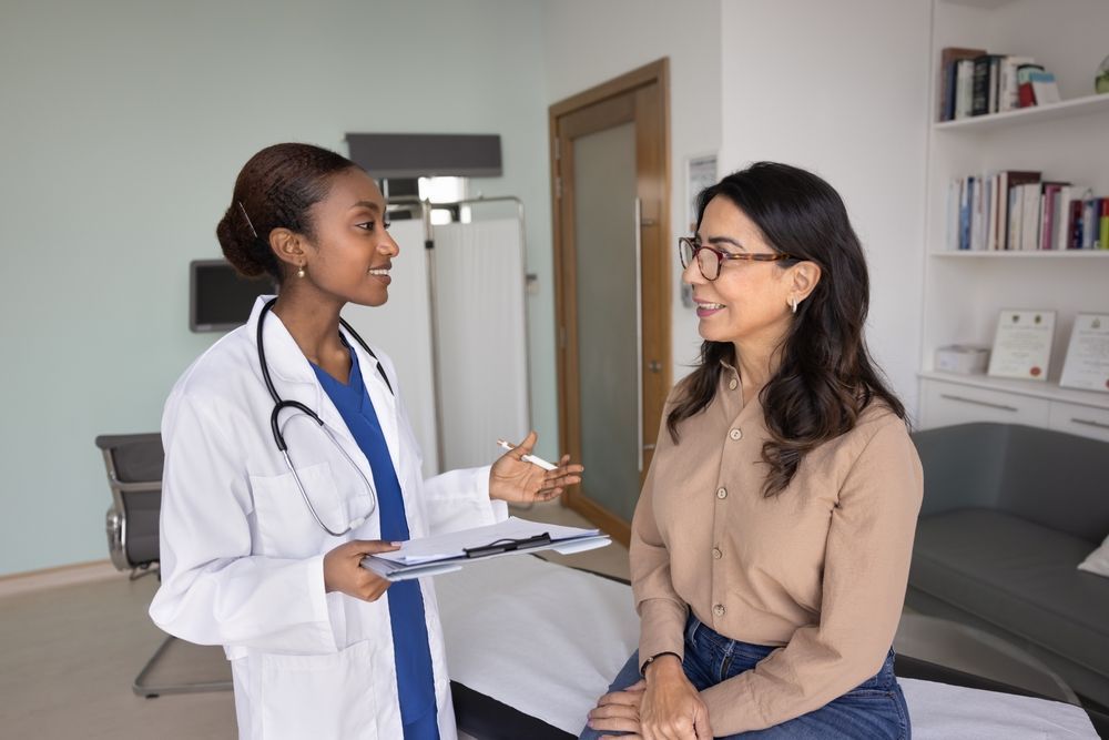 Doctor talking with seated patient in a clinic room, holding a clipboard.