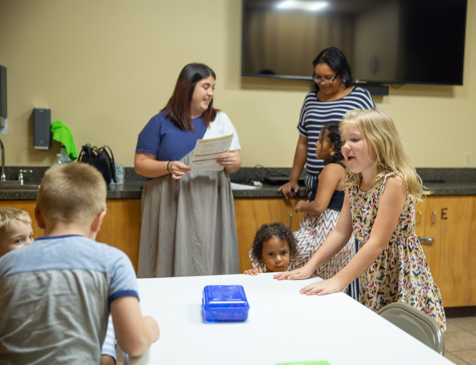 A group of children are sitting at a table with a woman standing behind them.