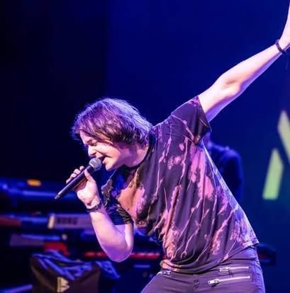 Zangeres treedt op het podium op met microfoon, arm omhoog, onder paarsblauwe concertverlichting.