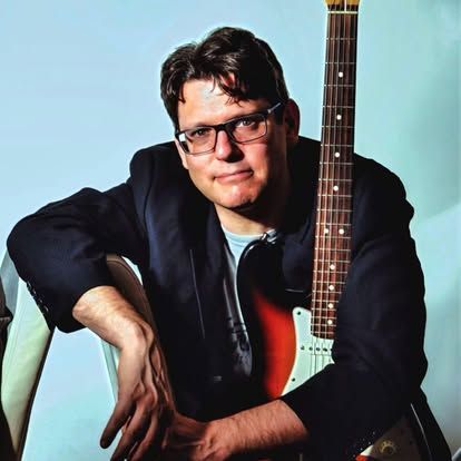 Portret van een man met een bril die tegen een elektrische gitaar leunt, tegen een blauwe achtergrond.