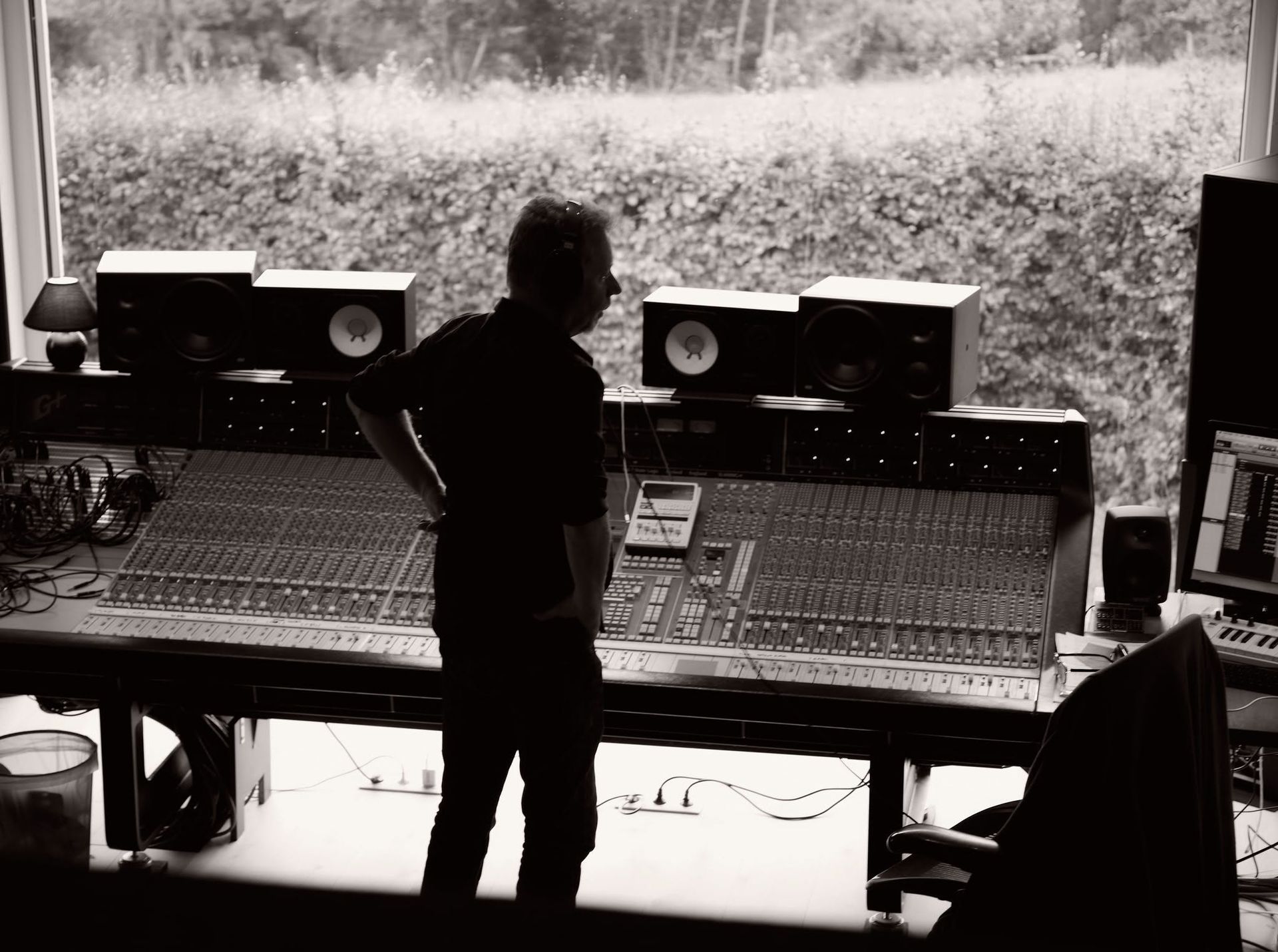 Een persoon in silhouet in een opnamestudio, met uitzicht op mengpanelen en luidsprekers, en een groot raam op de achtergrond.
