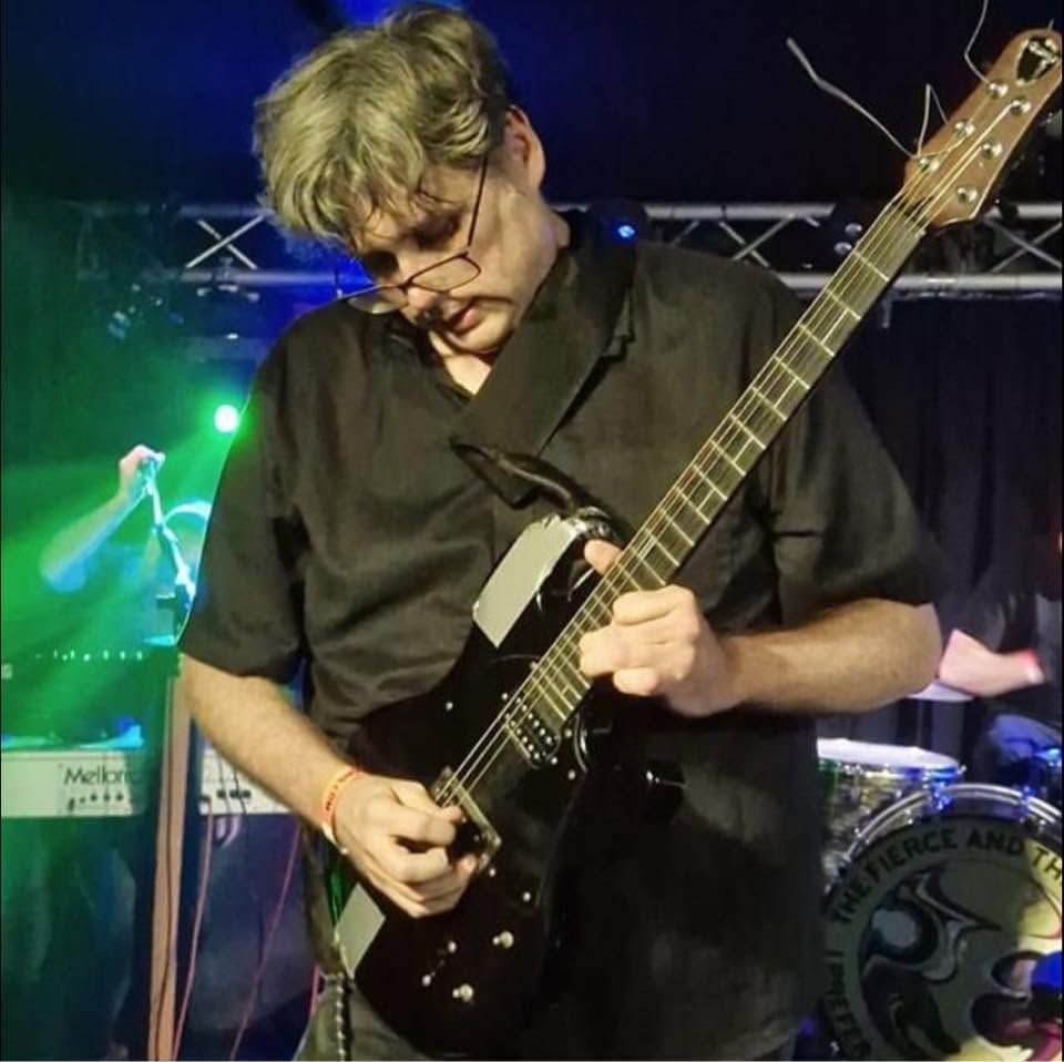 Gitarist in een zwart shirt die op een elektrische gitaar speelt op het podium onder groene lichten.