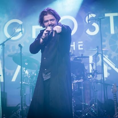 De zanger, gekleed in een lange zwarte jas, wijst naar het publiek op een blauwverlicht concertpodium.