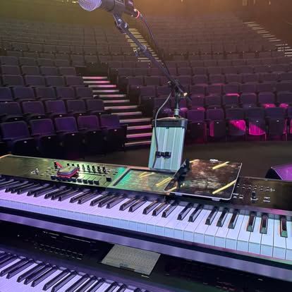 Keyboard op het podium in een lege zaal met microfoonstandaard en roze verlichte stoelen.