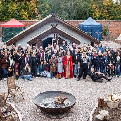 Een grote groep poseert buiten voor een blokhut, met een vuurplaats op de voorgrond en tenten aan weerszijden.