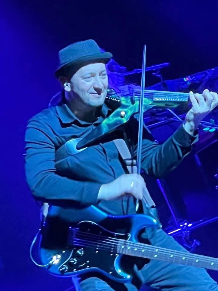 Een muzikant met een zwarte hoed speelt viool op een blauwverlicht podium, met een gitaar aan zijn riem.