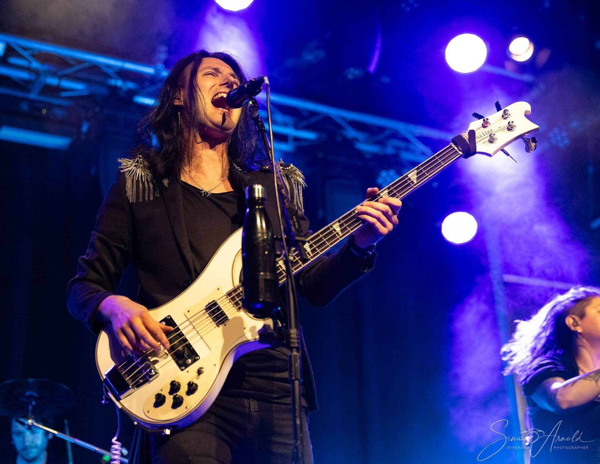 Zanger speelt op een witte basgitaar op een blauw verlicht concertpodium en zingt in een microfoon.