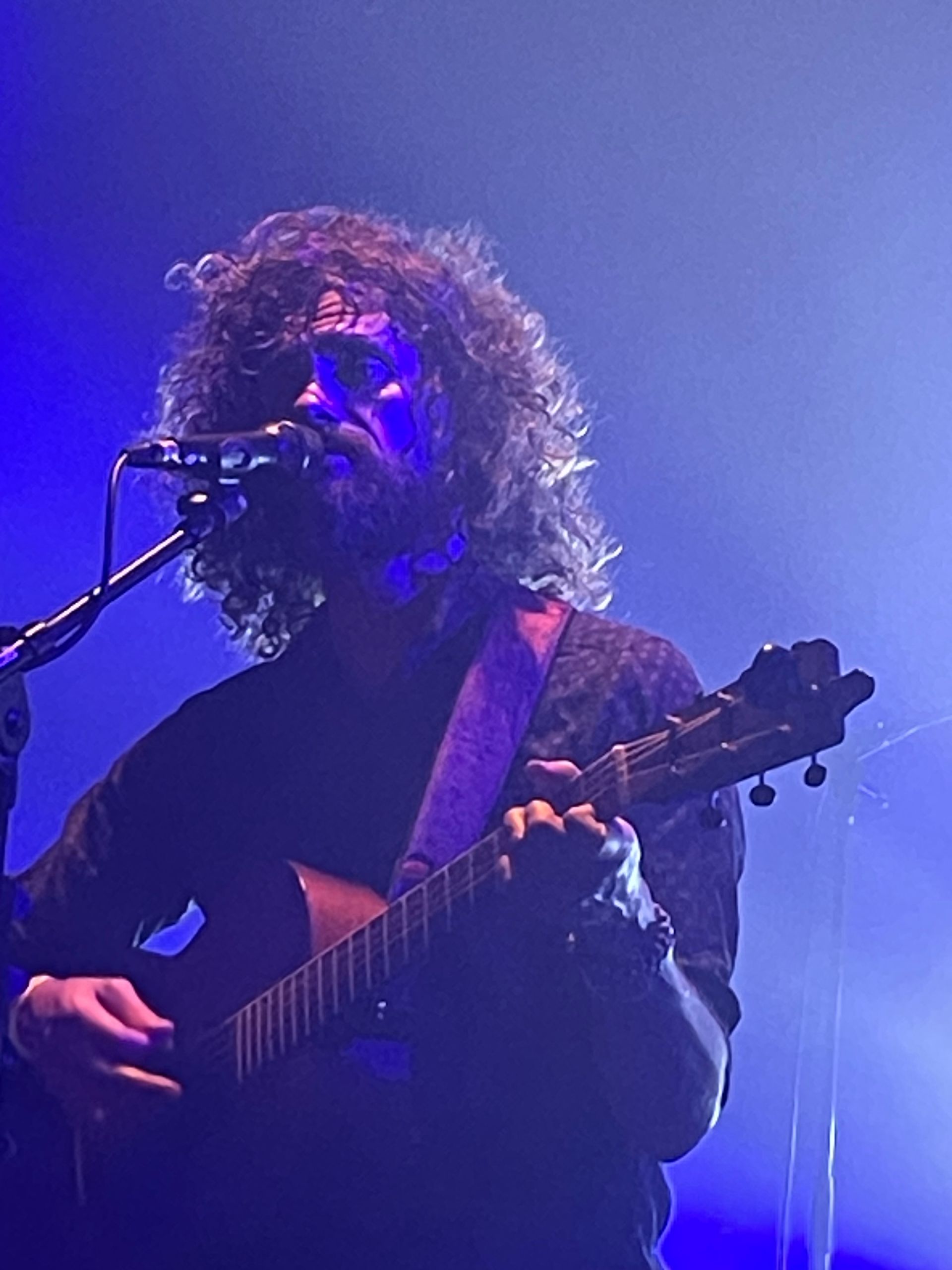 Zangeres met krullend haar die gitaar speelt en in een microfoon zingt onder blauwpaarse podiumverlichting.