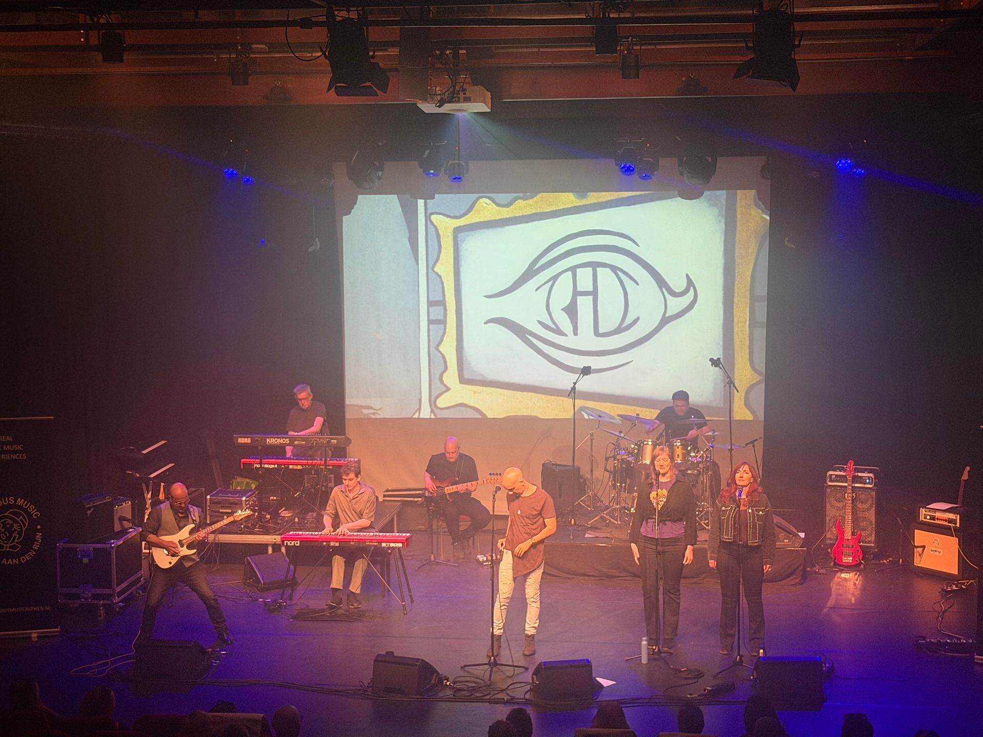 Een liveband treedt op op een schemerig verlicht podium, met een groot ooglogo dat op de achtergrond wordt geprojecteerd.
