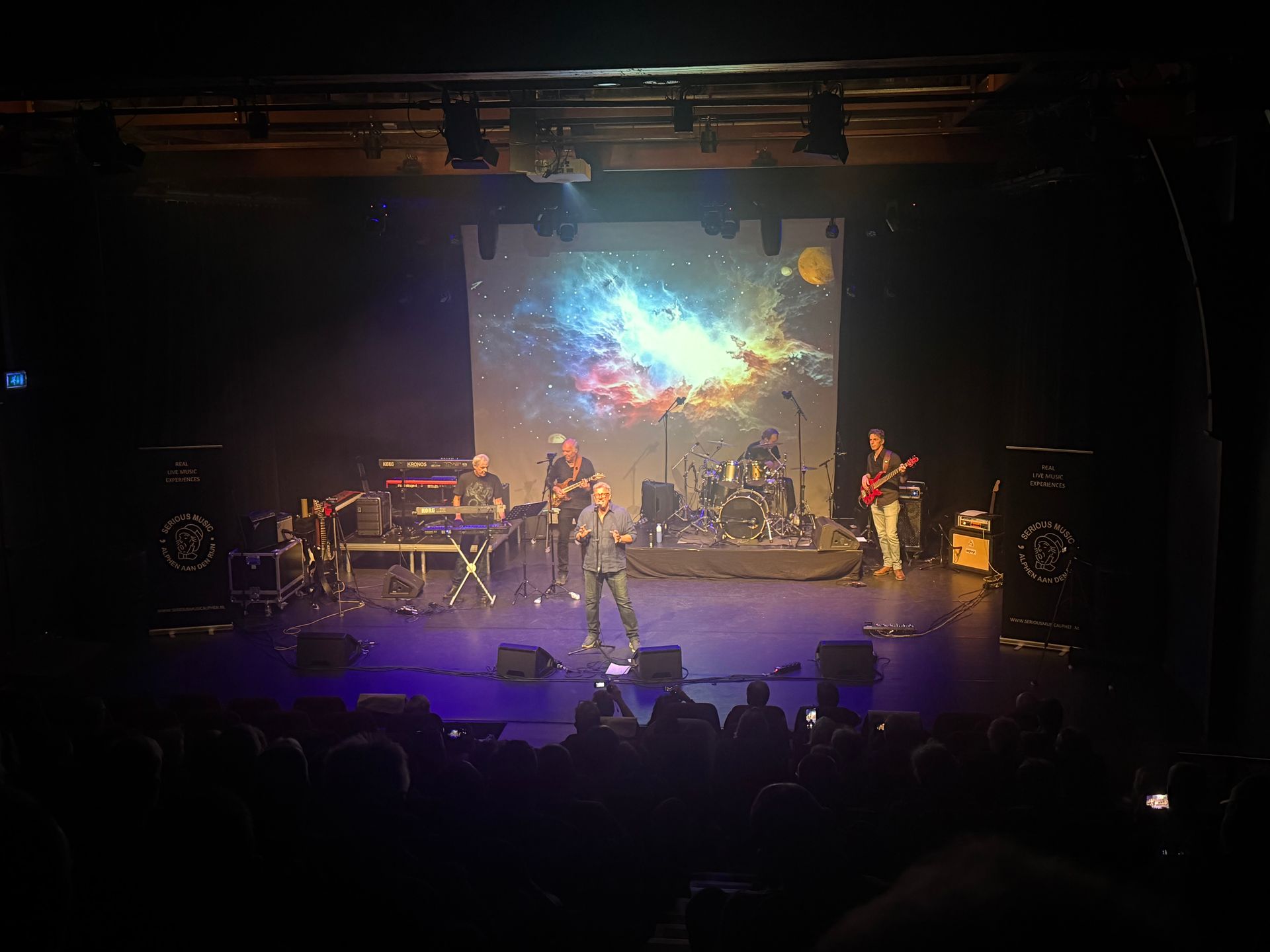 Een liveband treedt op op een schemerig verlicht podium, met kleurrijke beelden van een sterrenstelsel op een scherm achter hen.