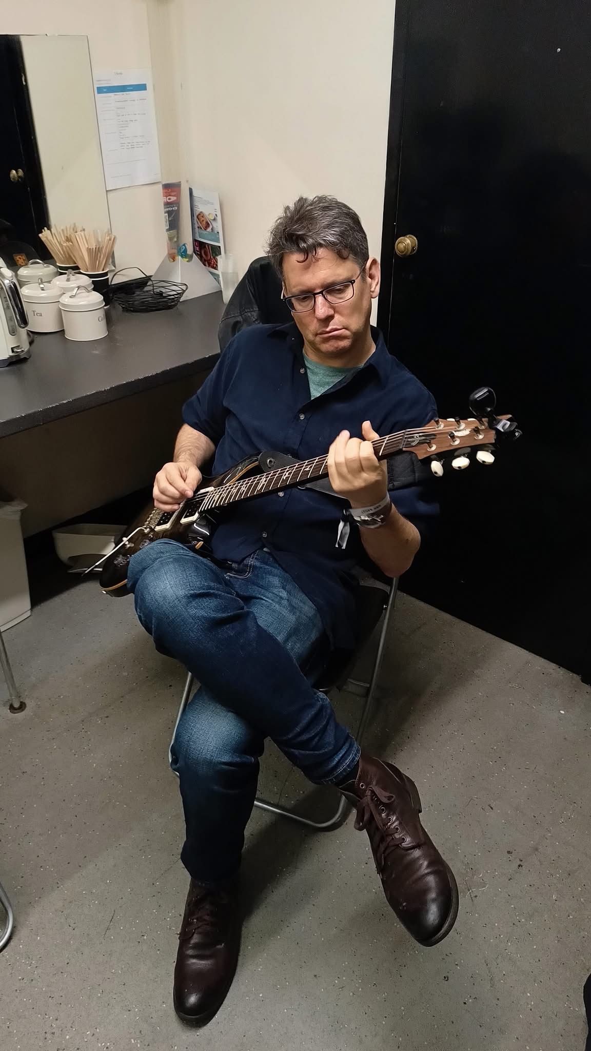 Een persoon zit in een kleine kamer elektrische gitaar te spelen, gekleed in een spijkerbroek en laarzen.