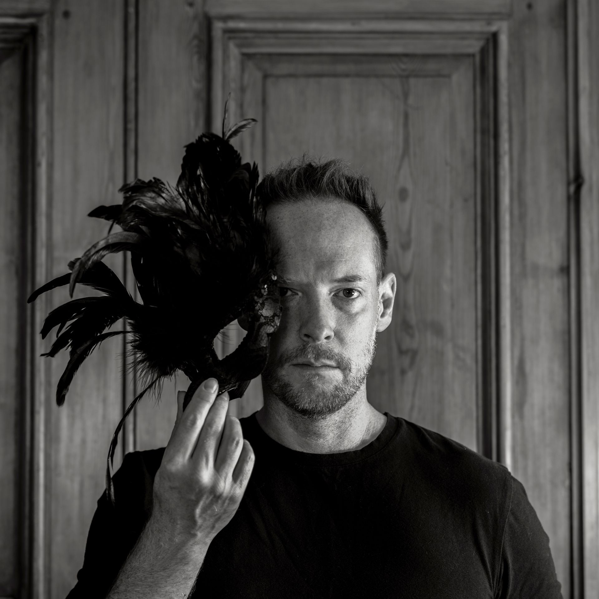 Zwart-wit portret van een man die een donker, gevederd masker voor één oog houdt, tegen een houten lambrisering.