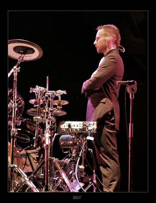 Een drummer staat naast zijn drumstel op een donker podium, met zijn armen over elkaar, onder roze verlichting.