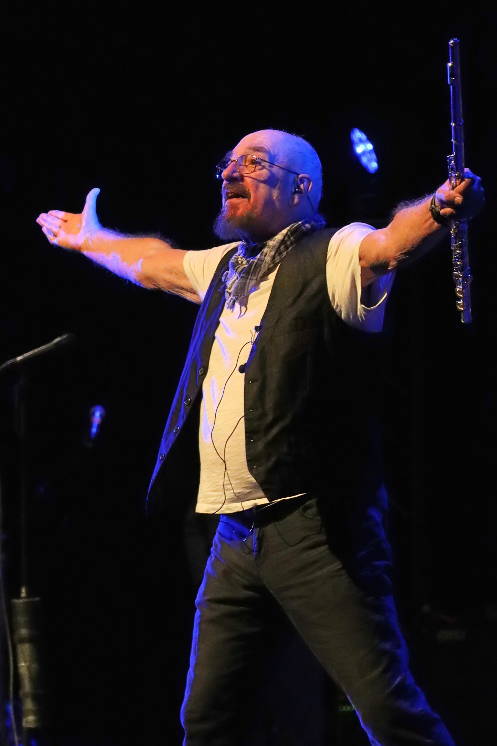 Man op het podium met uitgestrekte armen, gitaar en microfoon in zijn handen onder blauwpaarse concertverlichting.