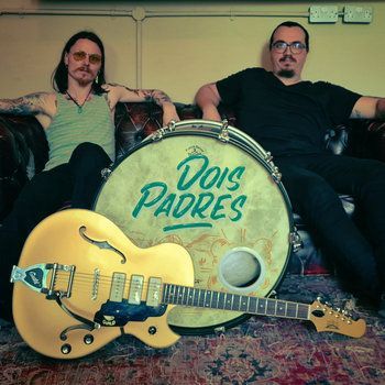 Twee mannen op een bank met een gele gitaar voor een grote trommel, waarop 