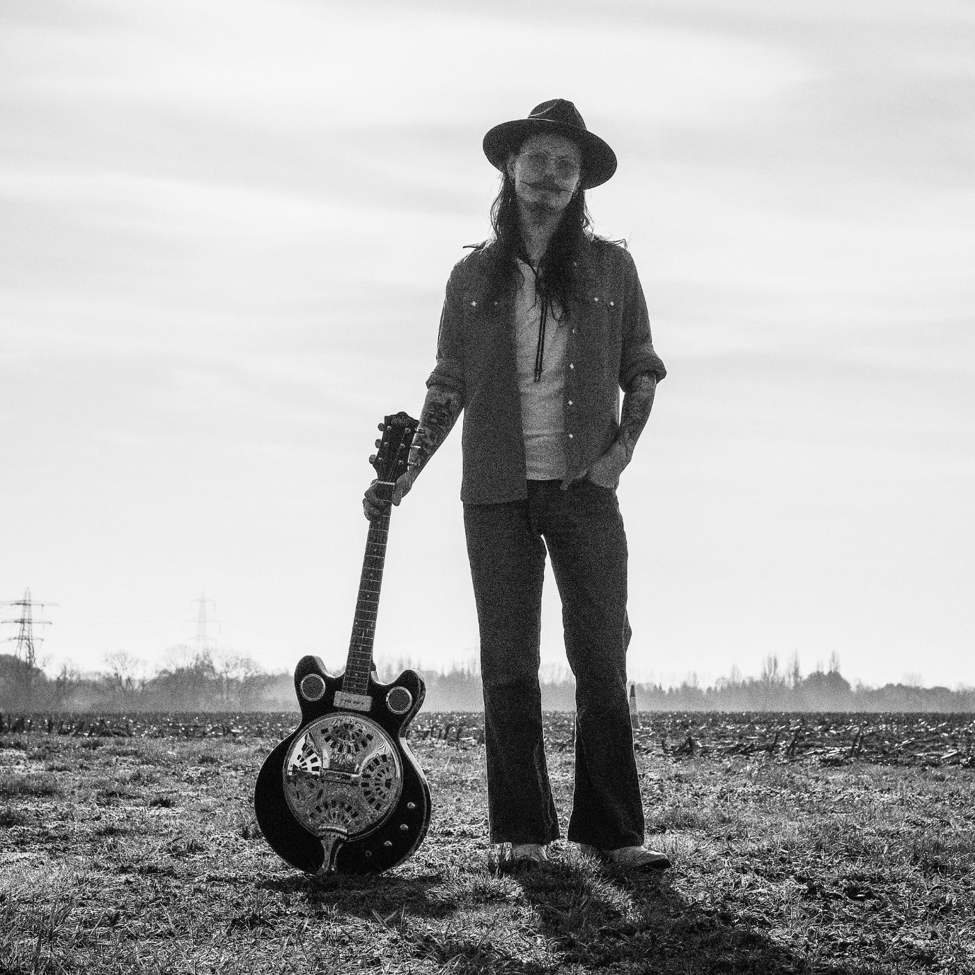Een persoon staat met een gitaar in een veld onder een bewolkte hemel.