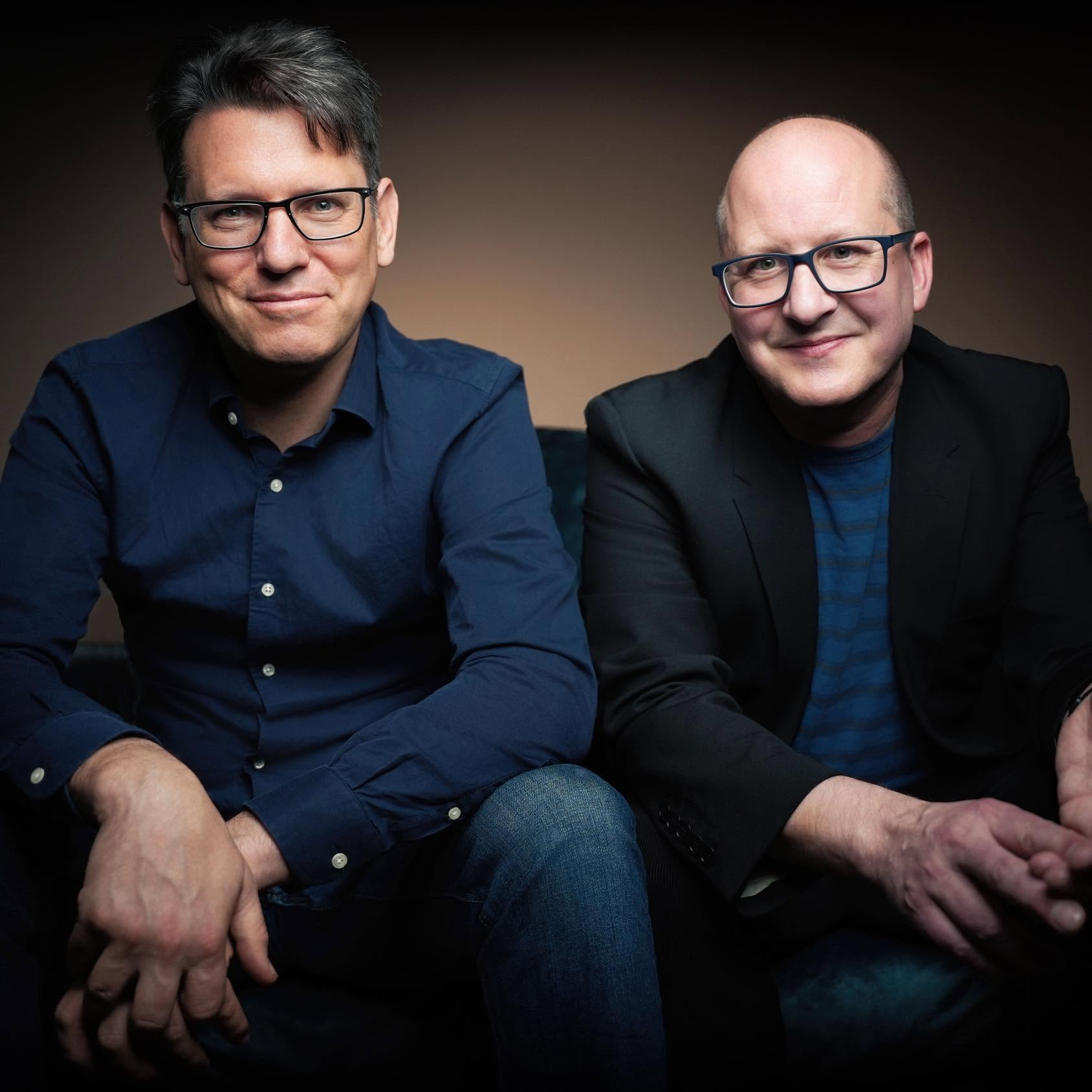 Twee mannen zitten naast elkaar in donkere jassen, tegen een donkere studioachtergrond, en kijken in de camera.